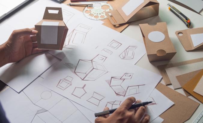 Person designing cardboard packaging and holding mockup for the design.”>
                  <div class=