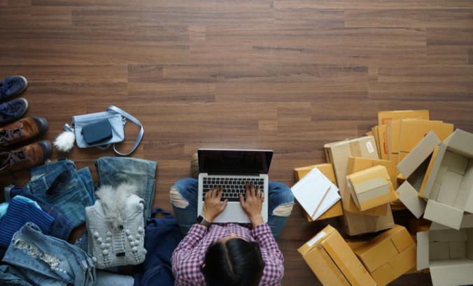 Person working laptop computer from home on wooden floor with postal parcels on right side and clothing and shoes on the left.