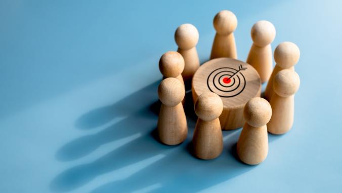 Target icon on round wood block surrounded with small wooden human figures